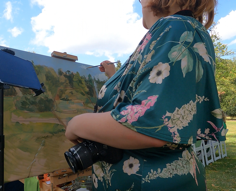 Foto Renée in groene bloemetjesjurk aan het schilderen. Buitenceremonie, op het doek schets van de achtergrond. Camera in de aanslag.