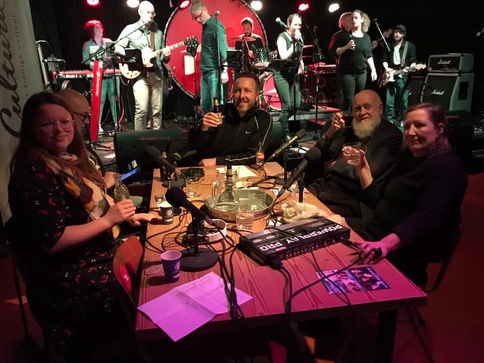 Foto van Renée en enkele andere gasten bij de lokale radio. Zittend aan tafel, allerlei apparatuur en drankjes erbij. Op de achtergrond een band op podium.