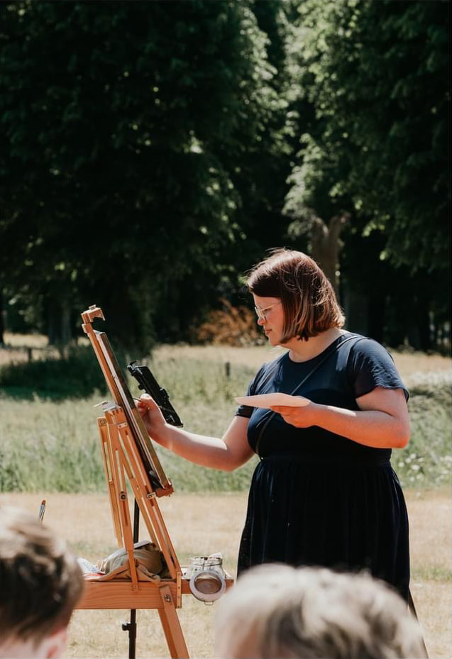 Outside photo, trees in the background. Wedding guests in the front. Renée is painting live on a painting on an easle. She is wearing a dark blue dress.