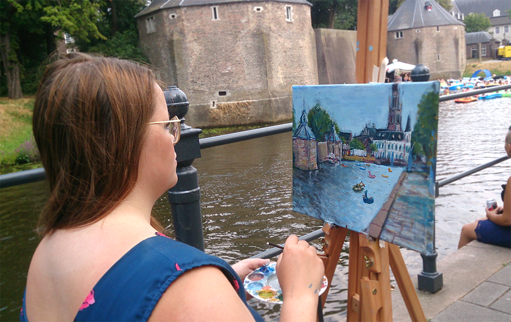 Foto van live painter Renée, aan de gracht in Breda, bij het spanjaardsgat. Op de schildersezel een doek van 30 bij 40 cm, met daarop het uitzicht op het water, de stoep en diverse gebouwen. In het water al enkele opblaasbootjes.