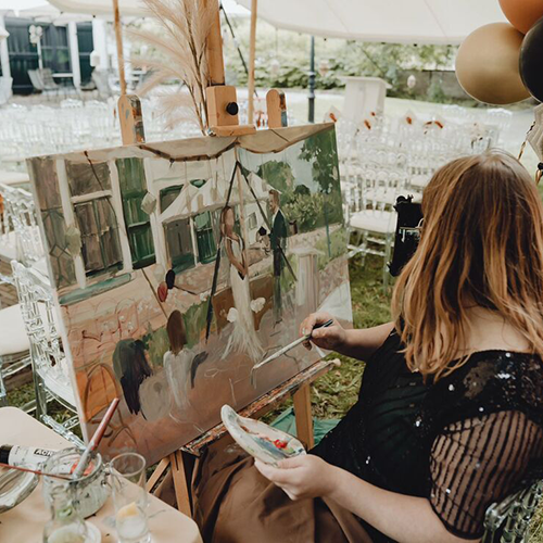 Foto van hoe ik aan het schilderen ben. Liggend doek met bruidspaar op schildersezel. Ik zit erbij en draag een zwart-groene flapper dress. Op achtergrond buitenlocatie met stoelen waar net de ceremonie was.