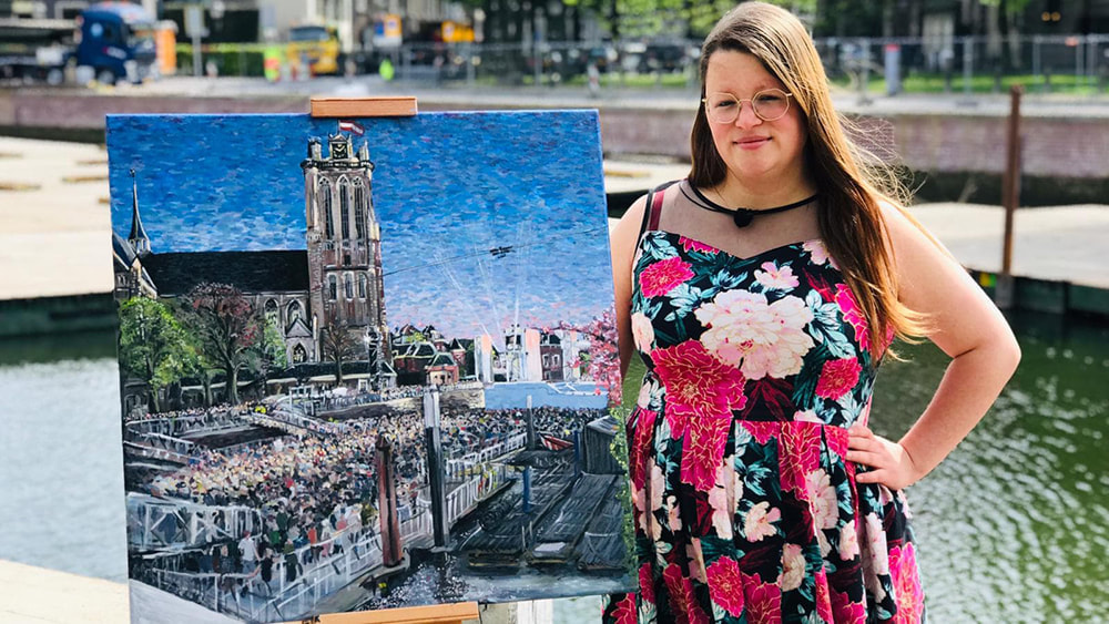 Foto Renée in roze bloemetjesjurk. Naast vierkant schilderij op schildersezel, aan de gracht. Schilderij is van The Passion in Dordrecht, uitzicht op mensenmassa en de grote kerk.