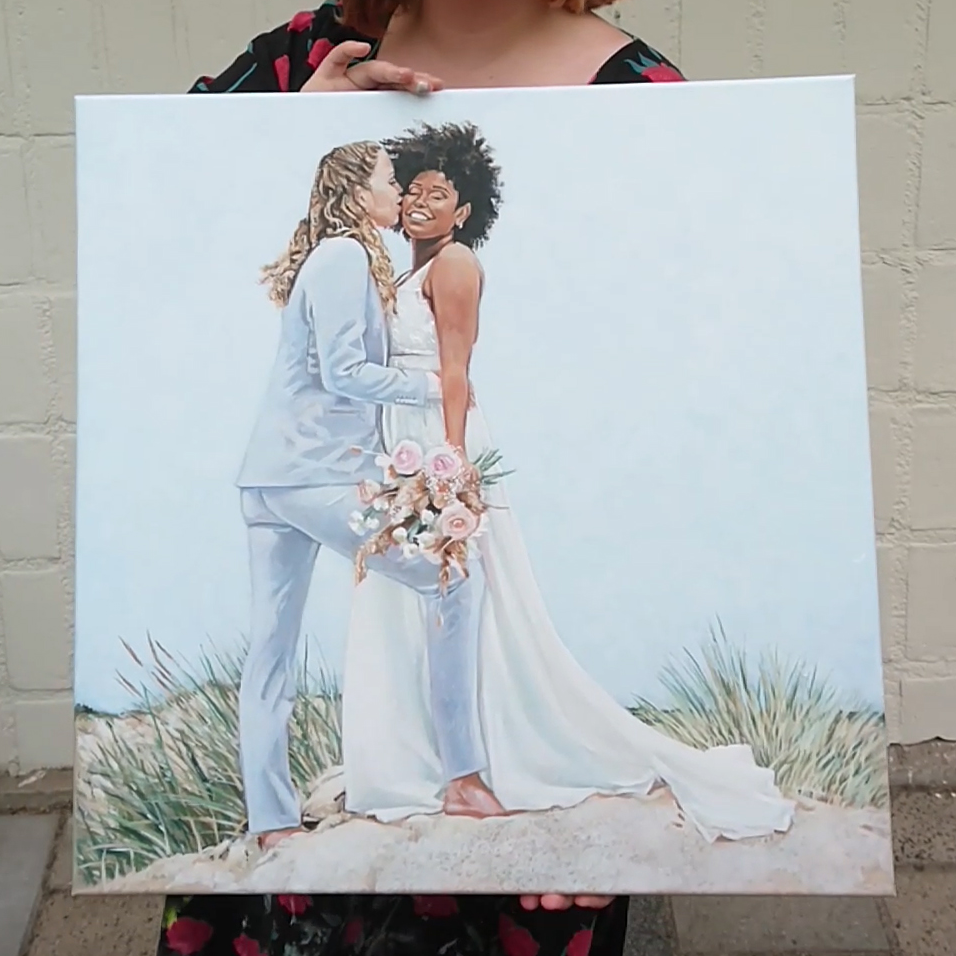 Holding a square canvas with a beach/dune setting. Bride in light blue suit kissing bride in white dress.