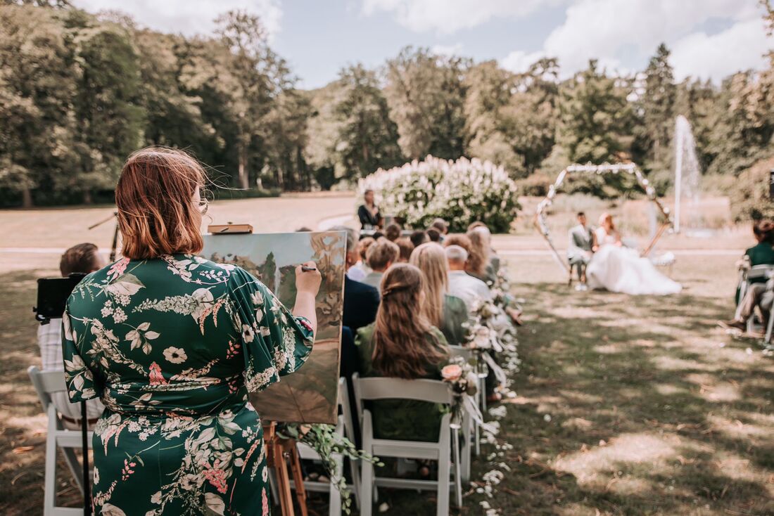 Photo of Renée live painting, standing behind sitting wedding guests. Looking to the sitting wedding couple in a big garden with a hexagon backdrop, a fountain, and lots of trees.